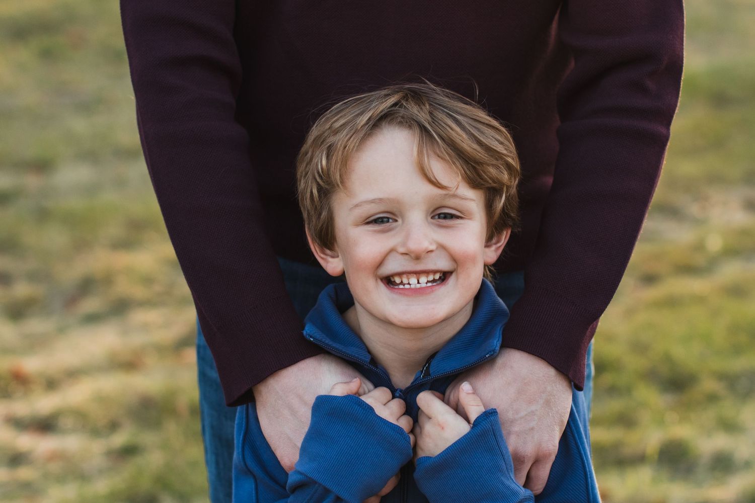 A cheerful young boy in a blue jacket grins while being hugged from behind by an adult outside.