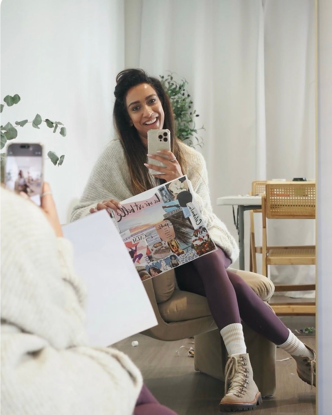 Smiling woman in cozy white sweater and purple leggings holds a magazine while taking a mirror selfie in a bright room.