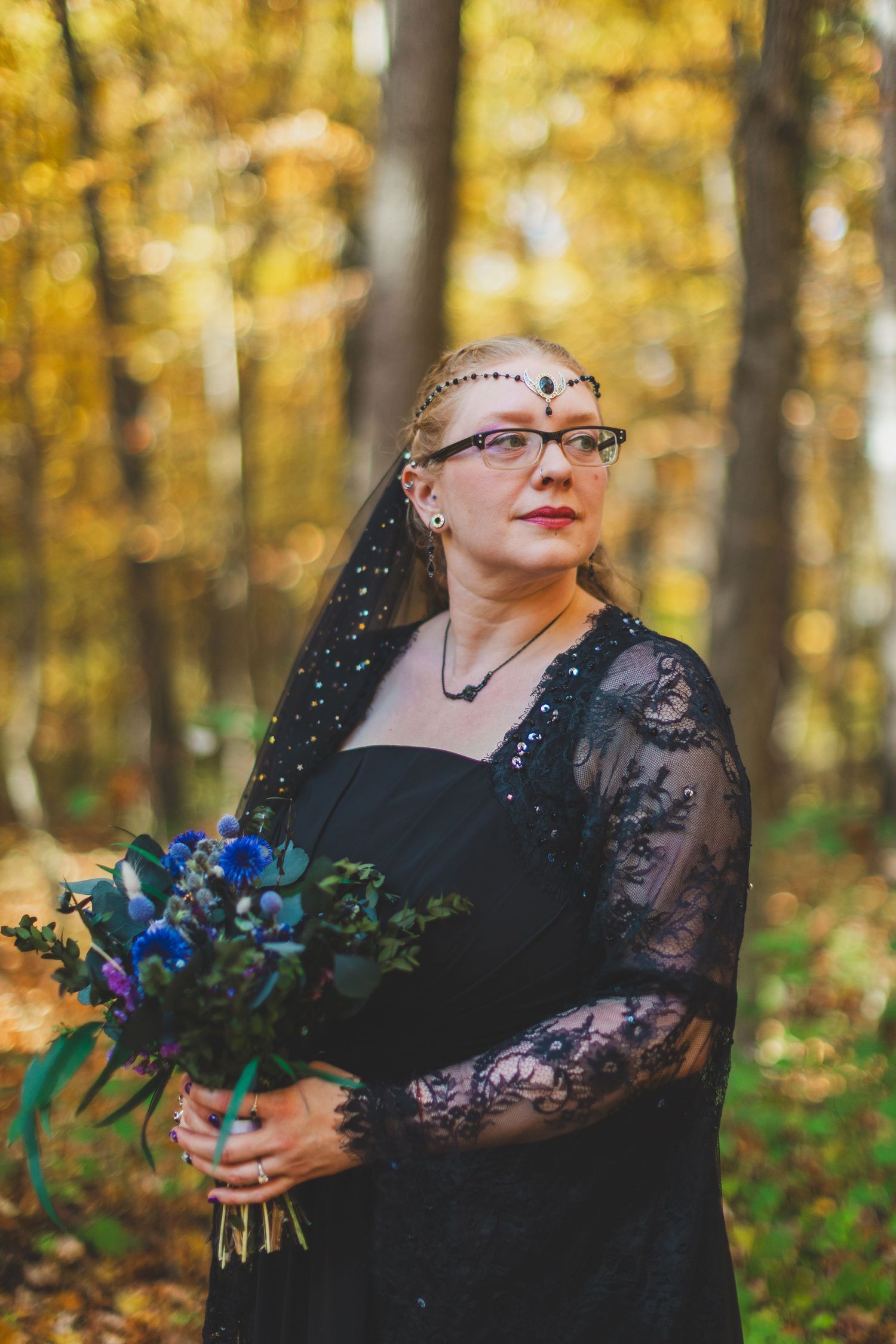 Woman in black lace dress and veil holding blue bouquet in autumn forest wedding portrait at Seneca Creek State Park