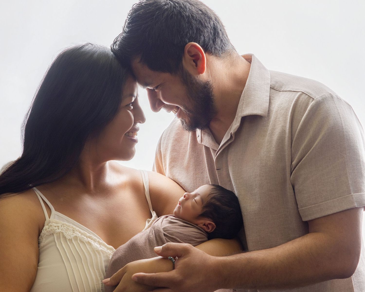 A couple tenderly holds their newborn baby, touching foreheads in an intimate family portrait against a soft white background.