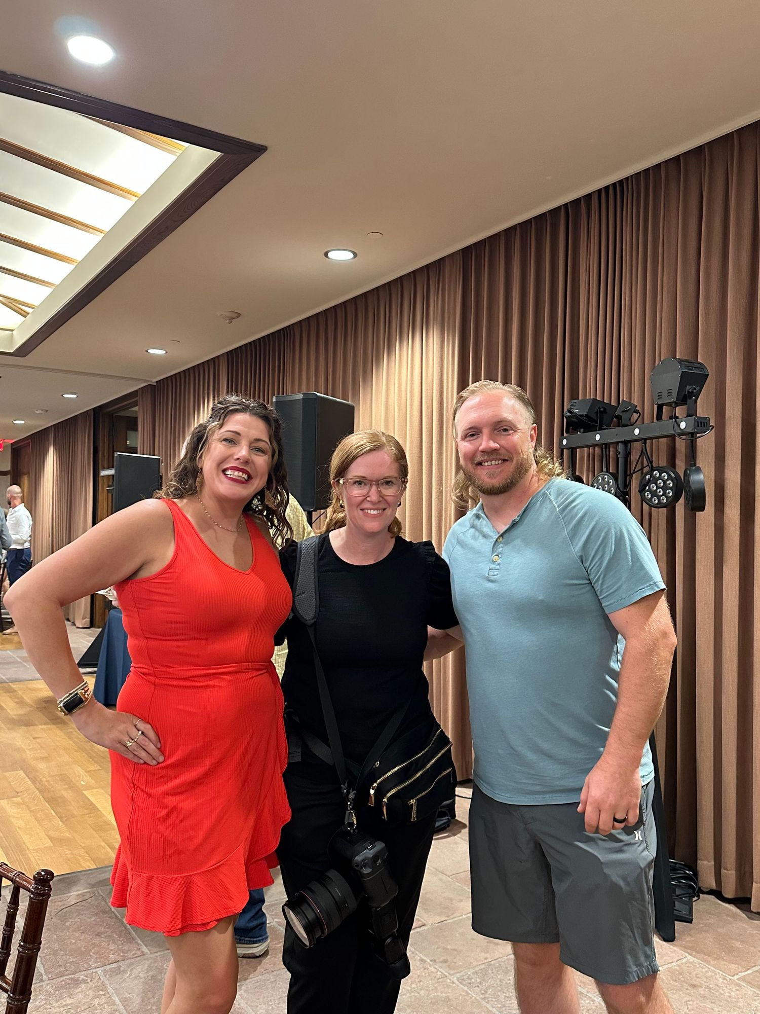 Three smiling people pose together in an elegant event venue with warm lighting and wooden curtains.