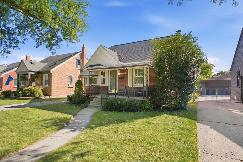 Charming brick bungalow with covered porch, lush green lawn, mature trees, and detached garage on a sunny residential street.