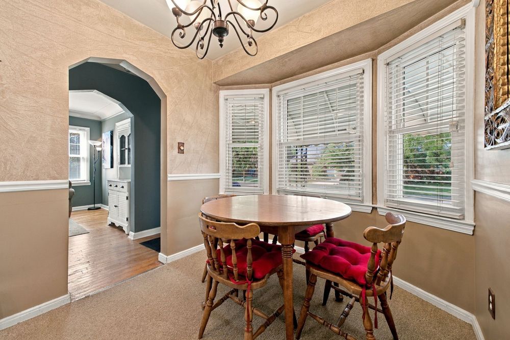 Cozy dining nook with round wood table, red cushioned chairs, bay windows with blinds, chandelier, and arched doorway.