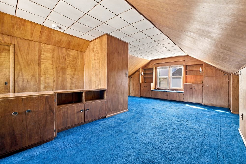 Attic room with wood-paneled walls, built-in cabinets, sloped ceiling, and bright blue carpet flooring.