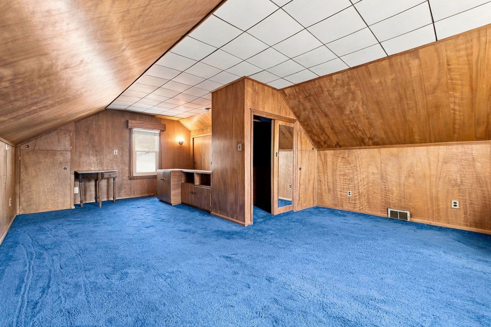 Spacious attic room with blue carpet, wood-paneled walls, angled ceiling, and built-in cabinetry in a residential home.