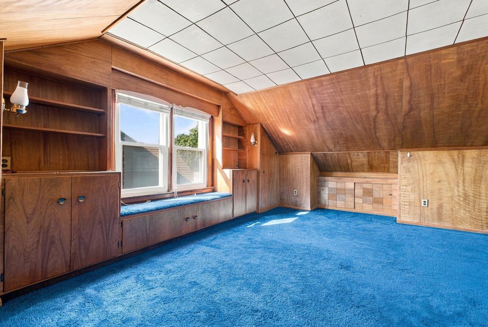 Attic room with blue carpet, wood-paneled walls, built-in shelves, cabinets, and a window with natural light.