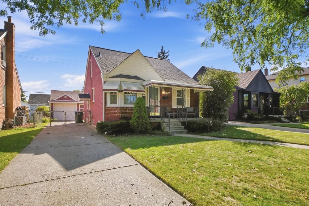 Charming brick bungalow with lush green lawn, mature trees, detached garage, and classic covered front porch on a sunny day.