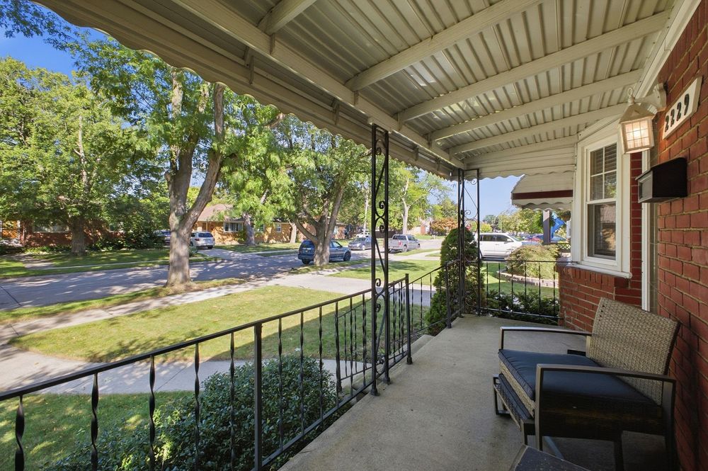 Covered front porch of a brick home with outdoor seating, black railings, and a view of a tree-lined residential street.