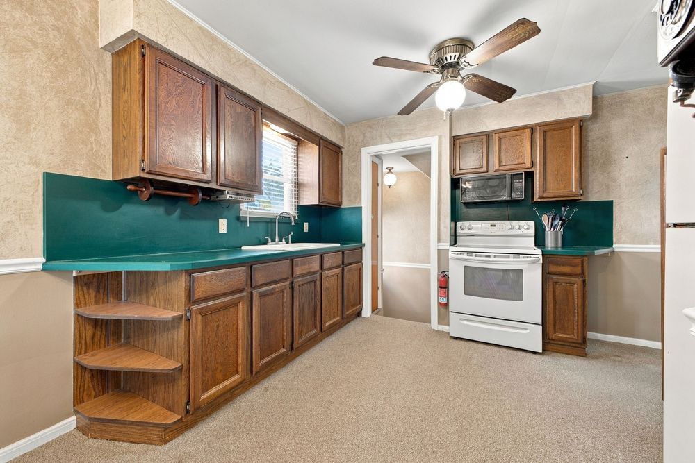 Kitchen with dark wood cabinets, green backsplash, white stove, microwave, ceiling fan, and carpeted flooring.