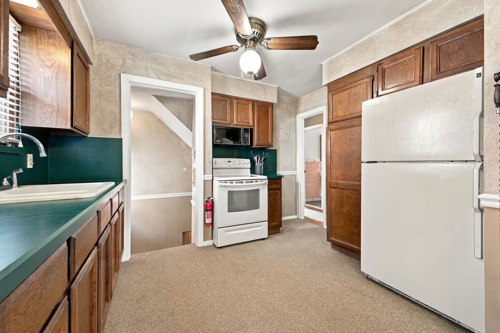 Kitchen with dark wood cabinets, green countertop, white appliances, and ceiling fan on carpeted floor.