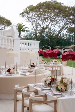Elegant outdoor wedding reception with cream chairs, floral centerpieces, and red parasols at a luxury venue.