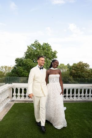 A bride and groom in elegant white wedding attire pose on a rooftop terrace with lush green trees in the background.