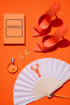 Orange flatlay with Hermès book, coral heels, hoop earrings, perfume, and a white fan with lobster print.