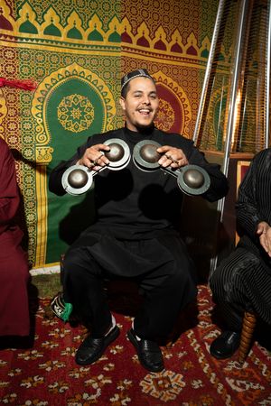 A smiling man in black attire plays traditional Moroccan krakeb castanets, seated before ornate Islamic patterned tapestries.