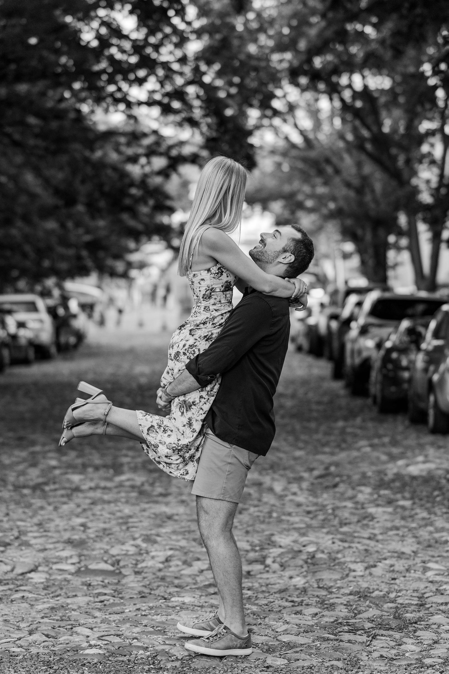 Black and white photo of a man lifting a woman in a floral dress on a cobblestone street lined with trees and cars.