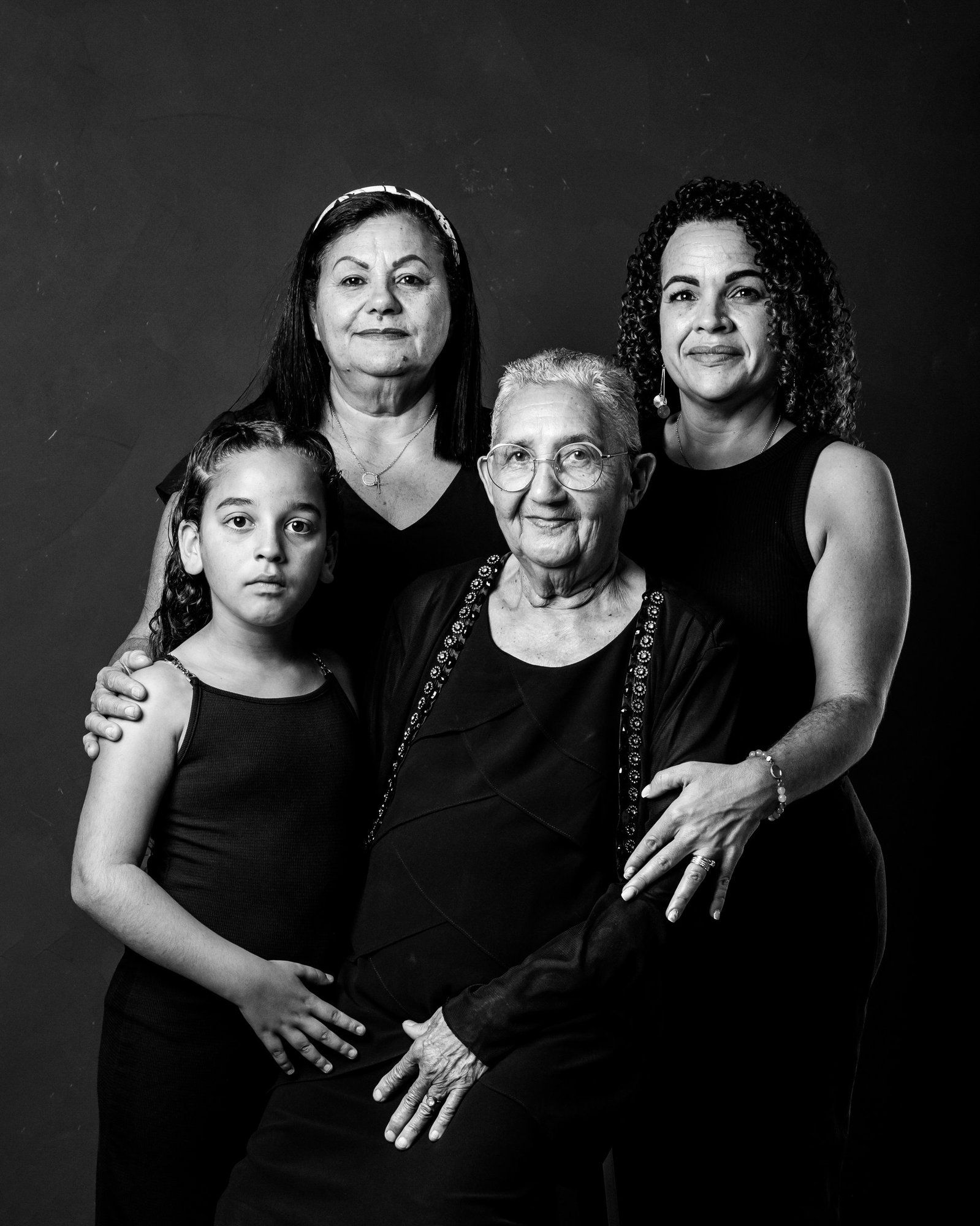 Black and white portrait of four generations of women posing together in matching black outfits against a dark background.