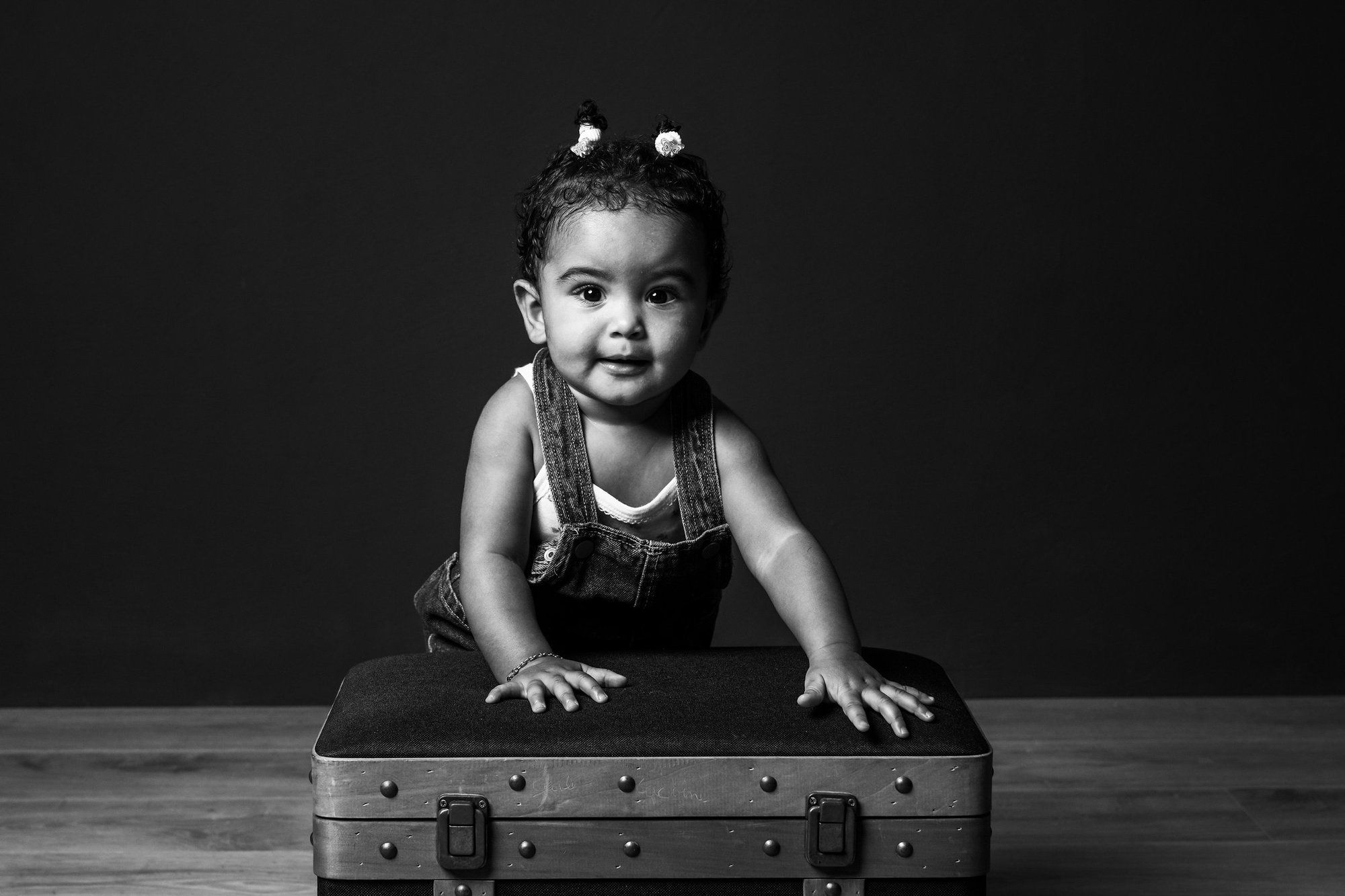 Black and white photo of a smiling toddler girl leaning on a vintage suitcase in a studio setting.