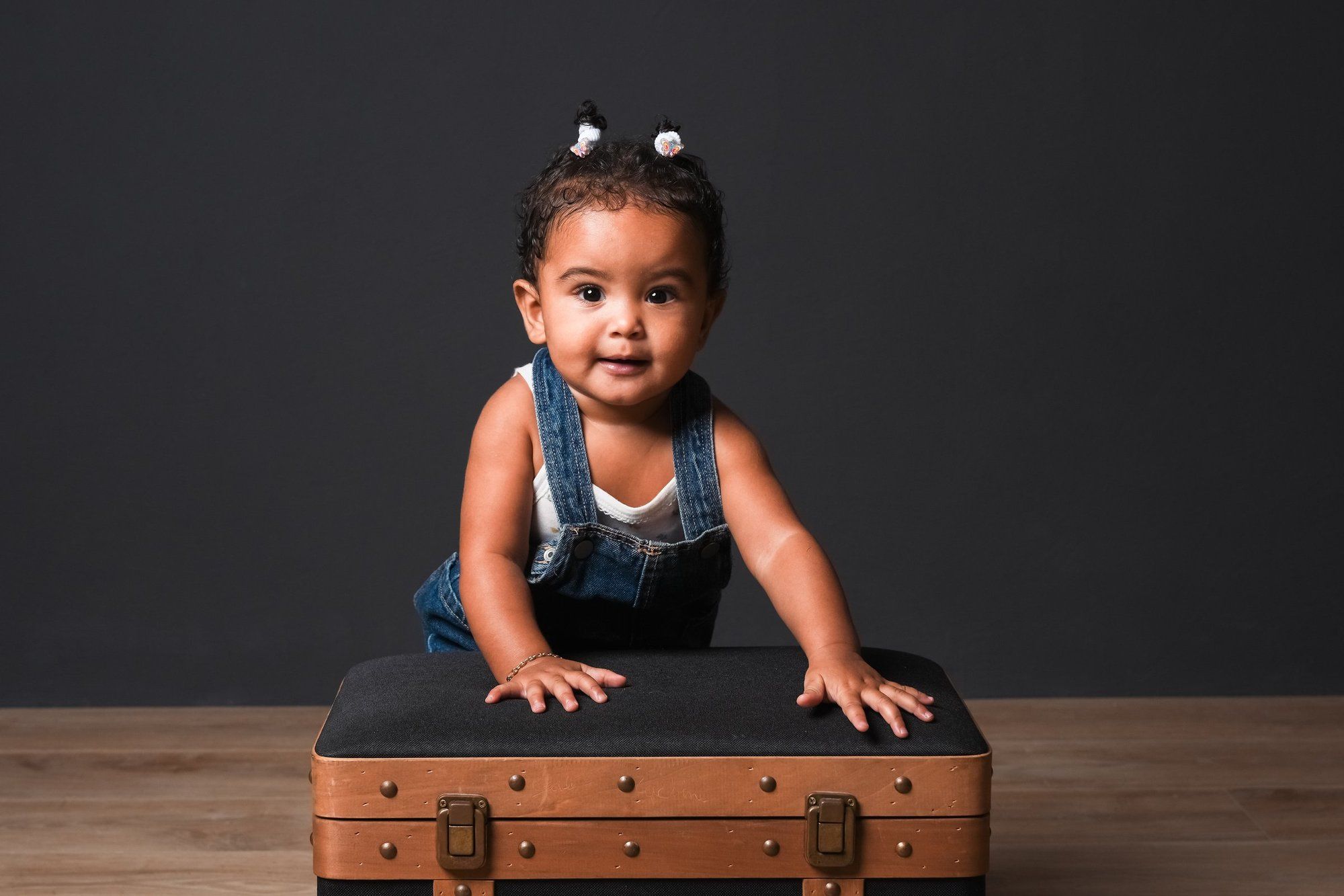 Adorable toddler girl in denim overalls leaning on a vintage brown suitcase against a gray background.