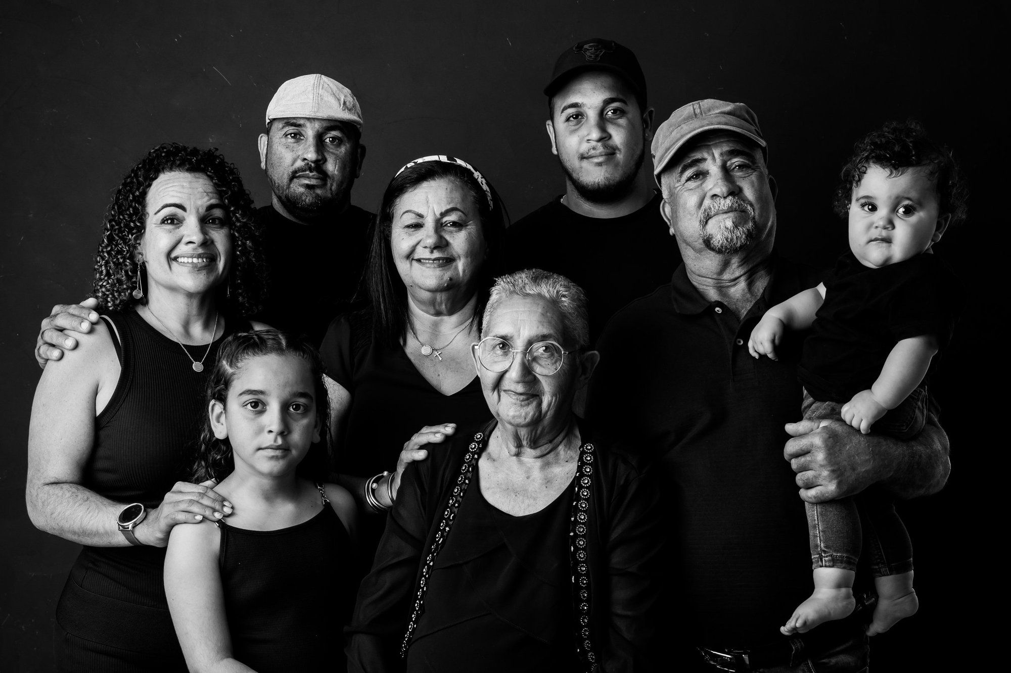 Black and white multigenerational family portrait with elderly woman centered, seven relatives posed together against dark background.