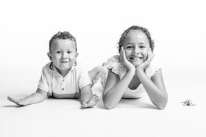 Black and white portrait of two smiling children lying on floor, boy in white shirt and girl with hands on cheeks.