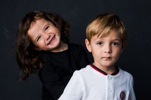 Two young children posing for portrait, smiling girl with dark hair hugging serious blonde boy against dark background.