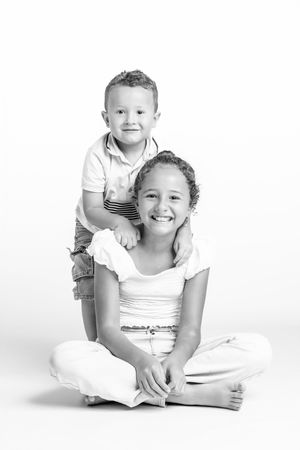Black and white photo of a smiling girl sitting cross-legged with a young boy standing behind her.