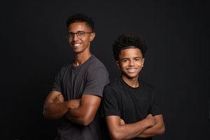 Two young men posing back-to-back with arms crossed, smiling against a dark studio background.