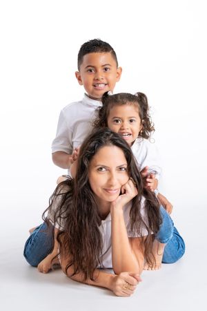 A smiling mother lies on the floor while her two young children stack playfully behind her in white shirts and jeans.