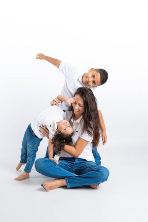 A mother sits cross-legged while her two children play around her, all wearing white tops and jeans.