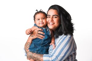 Smiling woman in striped shirt holding a happy toddler girl in denim dress against white background.