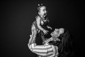 Black and white photo of a joyful mother lifting her laughing baby girl up in the air against dark background.