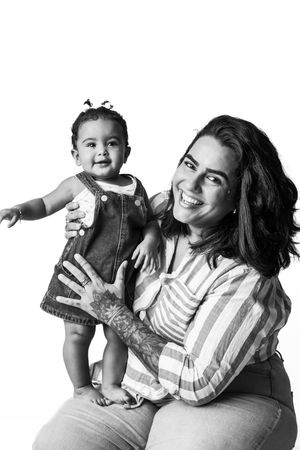 Black and white photo of a smiling woman holding a happy baby girl in a denim dress against white background.