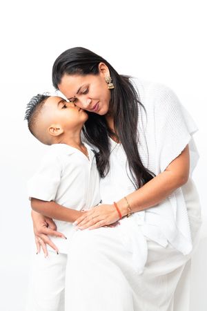 A young boy kisses his mother on the cheek as they share a tender moment, both dressed in white.