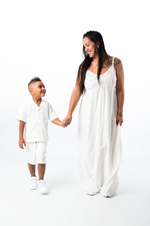 A smiling woman in a white maxi dress holds hands with a young boy in white outfit against a white background.