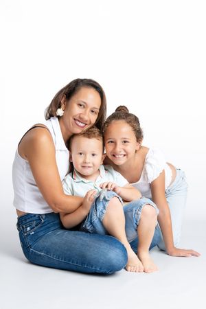 A smiling mother sits cross-legged with her two children, a toddler boy and older girl, posing together on white background.