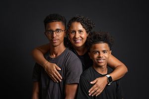 A smiling family of three in matching gray shirts pose together against a dark studio background.