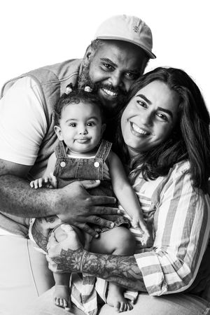 Black and white family portrait of smiling parents with tattooed father in cap holding their baby in overalls.