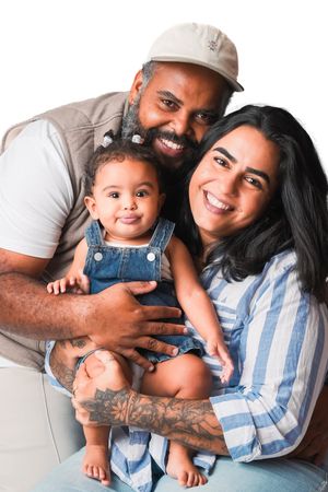 Smiling family portrait with tattooed father in cap, mother with dark hair, and baby in denim overalls on white background.