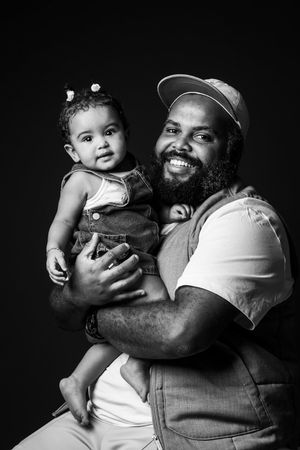 Black and white portrait of a bearded man in a cap holding a smiling baby girl in denim overalls.
