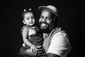 Black and white portrait of a smiling man holding a baby girl in overalls against a dark background.