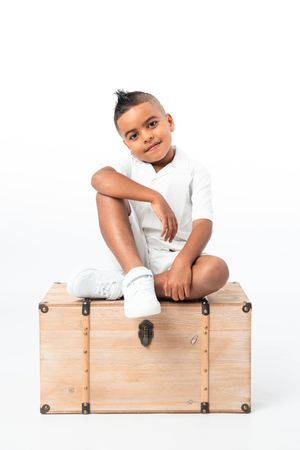 Young African American boy in white outfit sitting cross-legged on a wooden chest against white background.