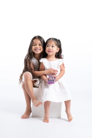 Two young girls in white dresses sit together smiling on a white cube, one holding a small purple box.