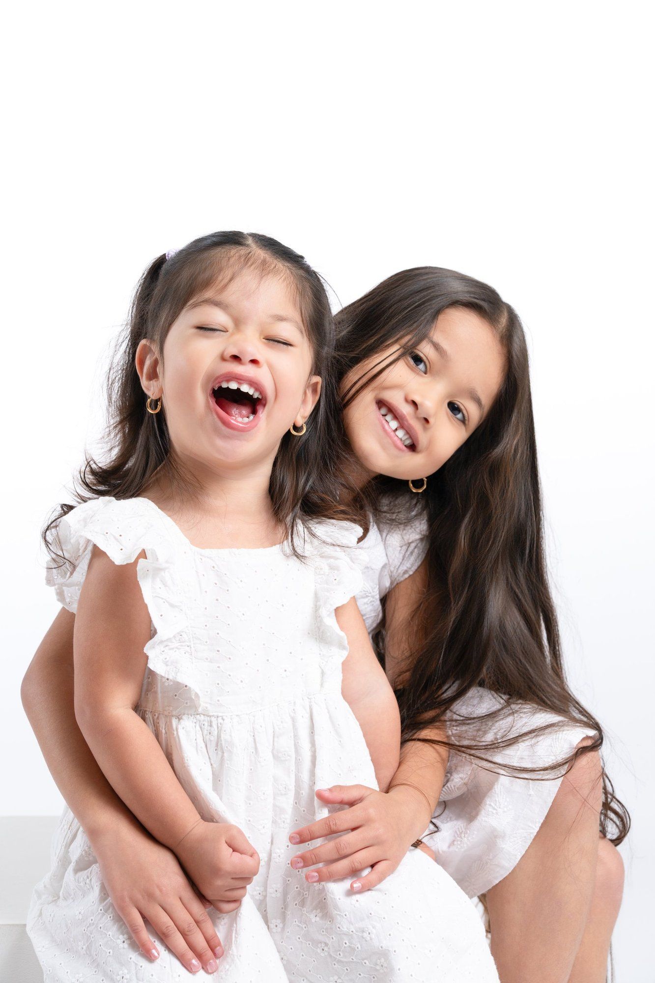 Two young girls in white dresses laughing and hugging together against a white background.