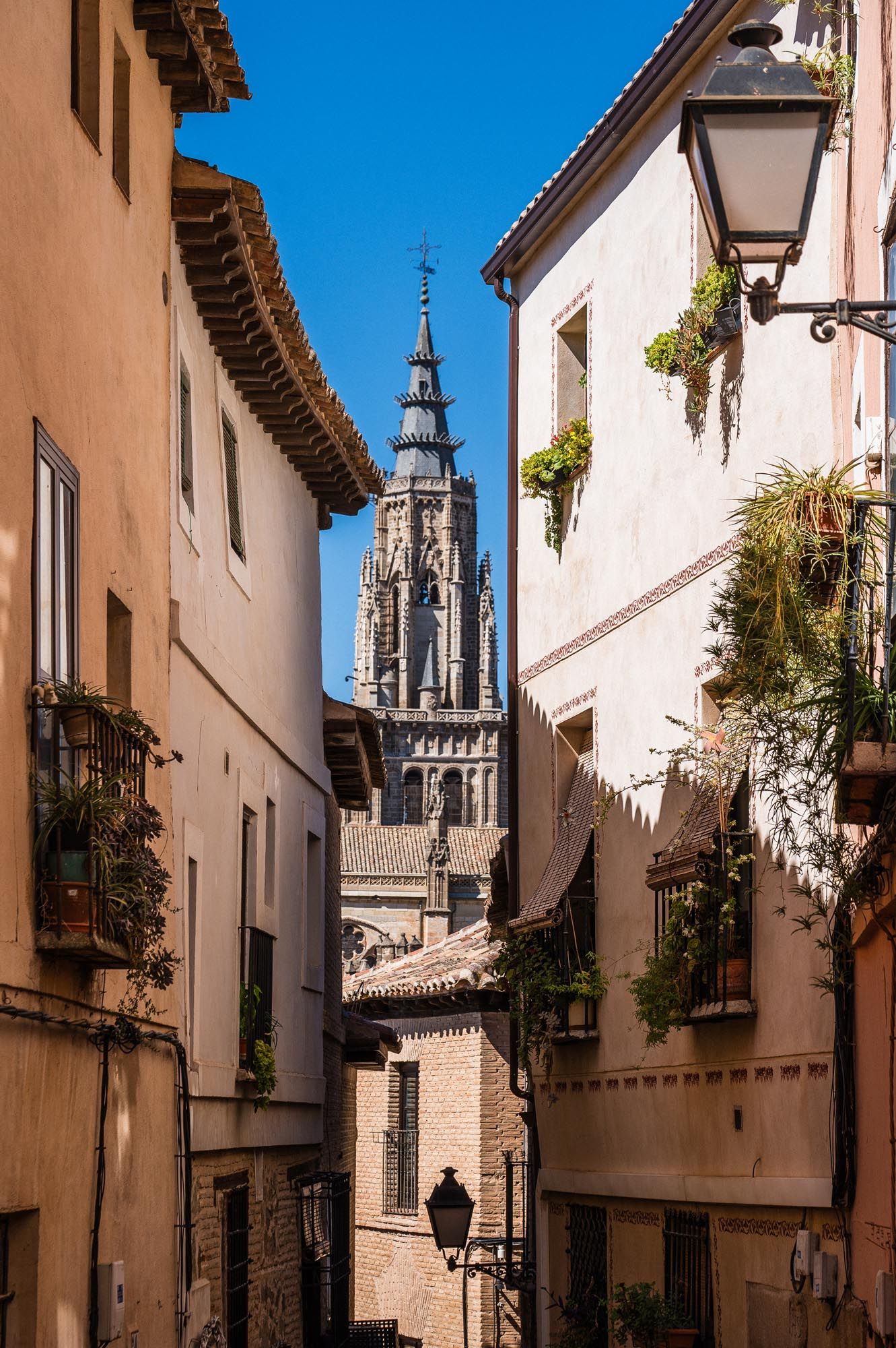 Narrow medieval street in Toledo, Spain with Gothic cathedral tower rising above historic sandstone buildings under blue sky.