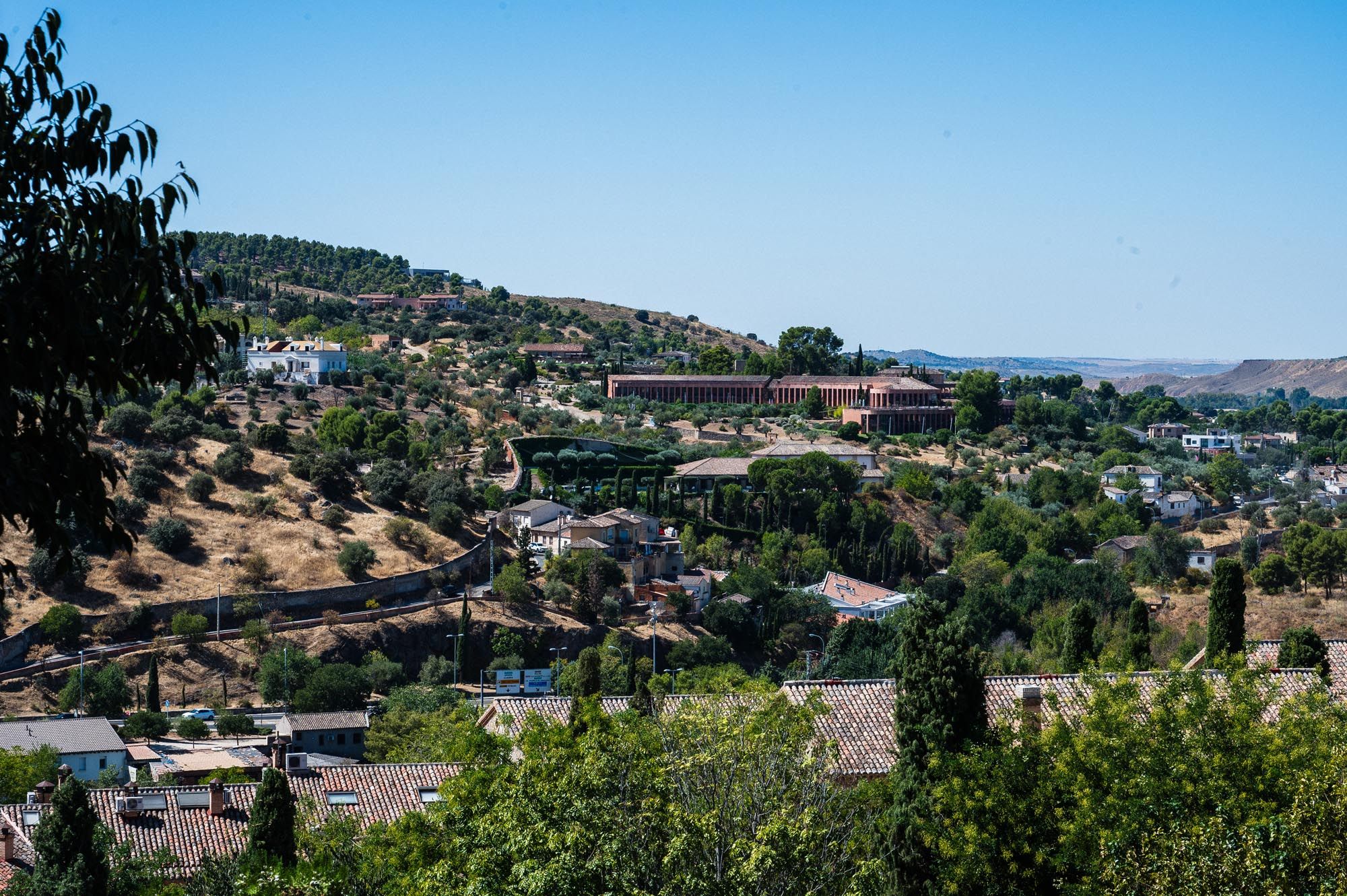 Aerial view of a hillside town with lush trees, buildings, and a winding road under a clear blue sky.