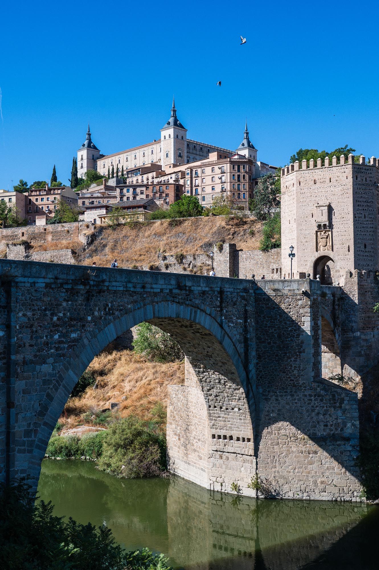 Historic Alcazar of Toledo rising above ancient stone bridge over Tagus River, Spain, under clear blue sky.