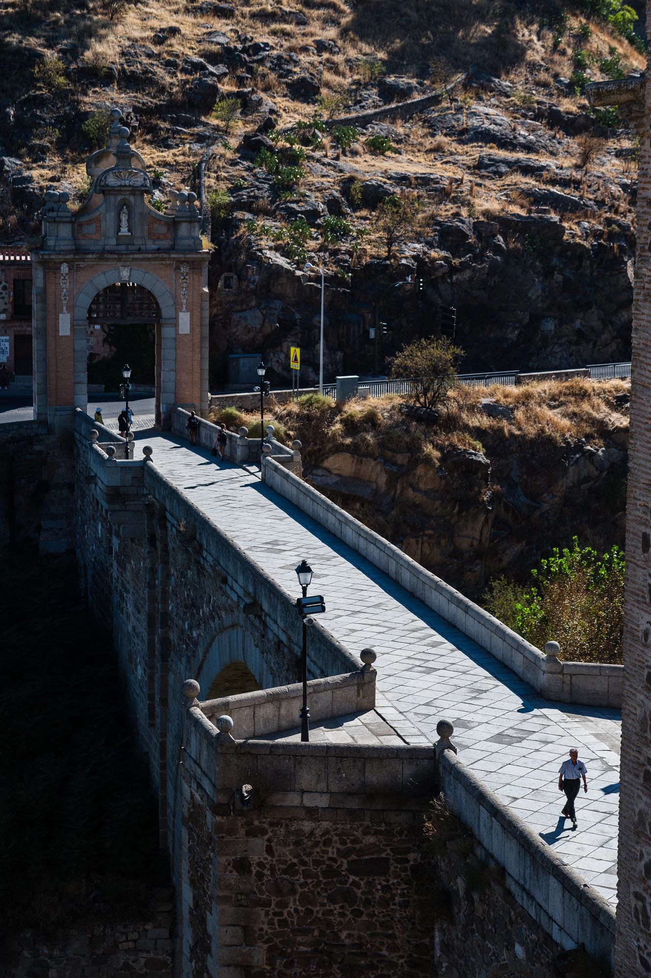 Historic stone bridge with ornate gate arch in Toledo, Spain, with a lone pedestrian walking under sunny skies.