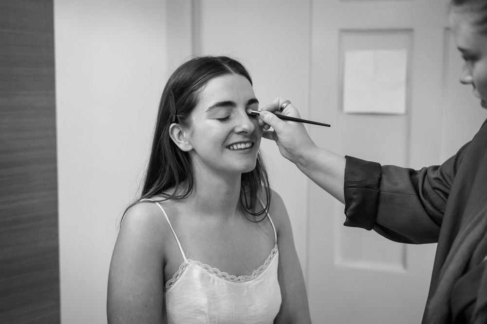 Black and white photo of a smiling woman getting eye makeup applied by a makeup artist.