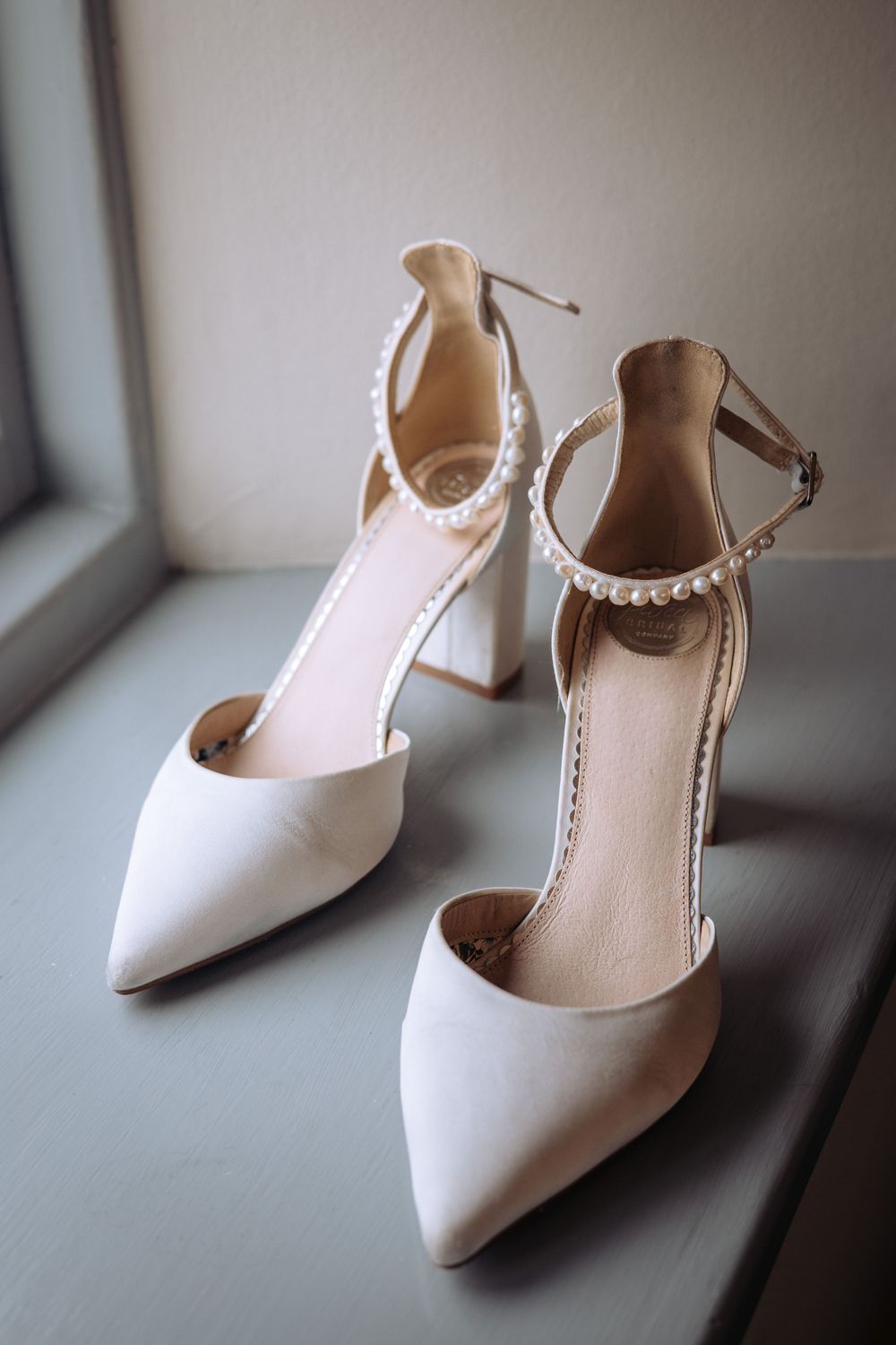 Elegant white block heel wedding shoes with pearl ankle strap detail displayed on a windowsill.