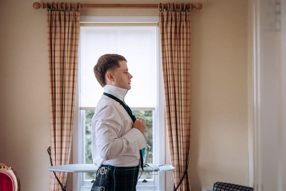 Groom in white shirt and kilt adjusting suspenders by a plaid curtained window while getting ready for wedding.